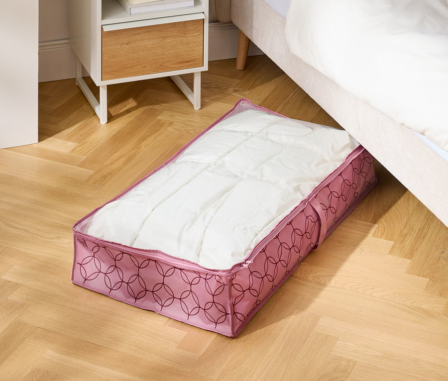 Auf einem Holzboden liegt eine rosafarbene Aufbewahrungstasche unter einem Bett neben einem Nachttisch.