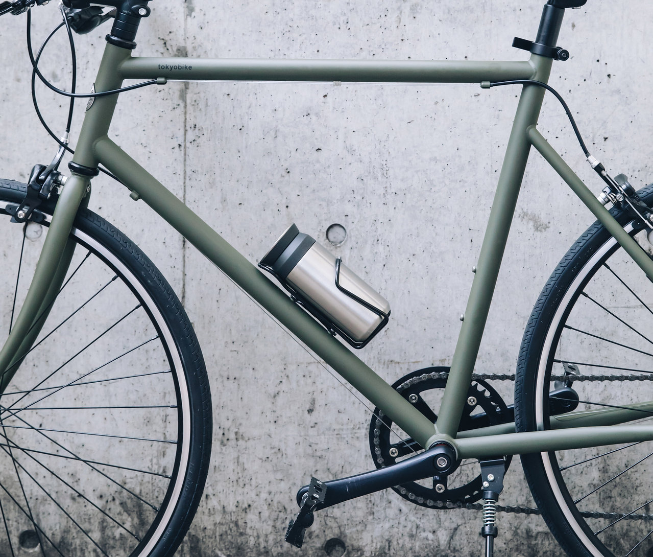 Nahaufnahme eines grünen Tokyobike Fahrrads an einer Betonwand, mit einer silbernen Wasserflasche in einem schwarzen Halter am Rahmen.