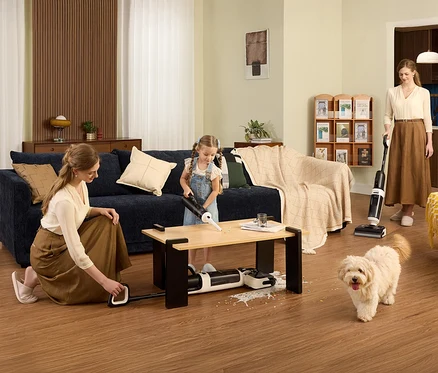 Eine Frau kniet und bedient den Tineco Nass-Trockensauger »Floor One Switch i5 Stretch«, während ein Mädchen einen Tisch saugt und eine andere Frau im Hintergrund staubsaugt.