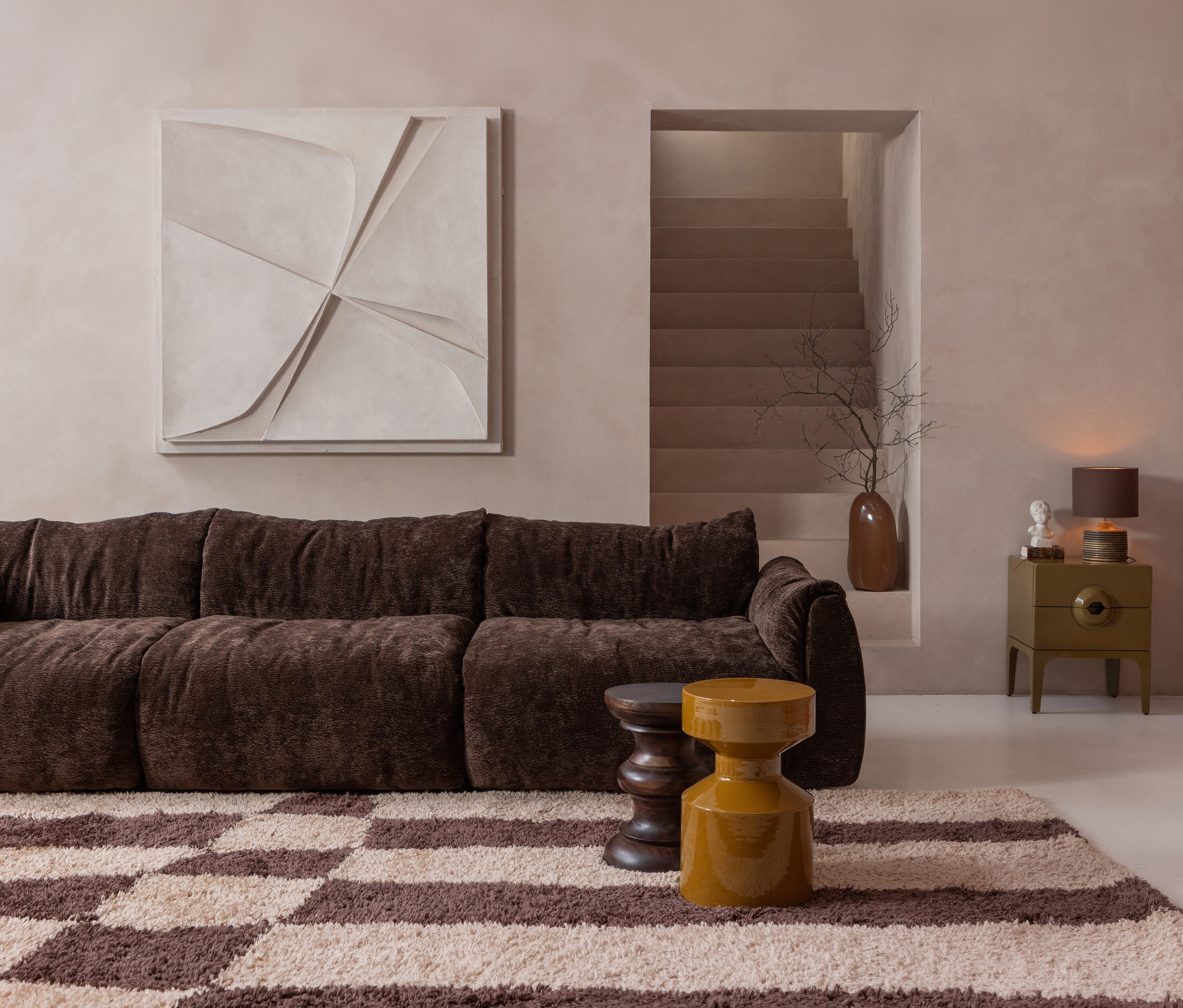Wohnzimmer mit braunem Sofa, Schachbrettteppich und gelber WOOOD »Merge« Kommode neben einer Treppe.
