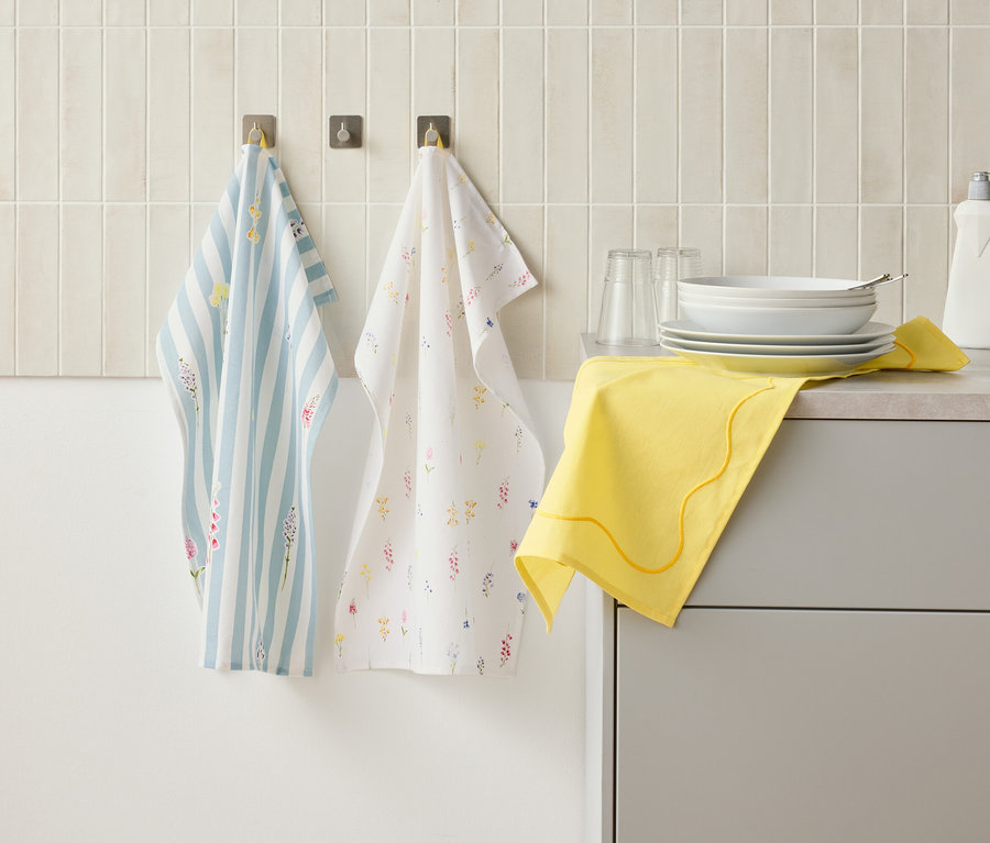 Zwei Geschirrtücher hängen an Haken an einer Wand über einer Küchentheke, auf der sich Geschirr, Gläser, eine Spülmittelflasche und ein gelbes Tuch befinden.