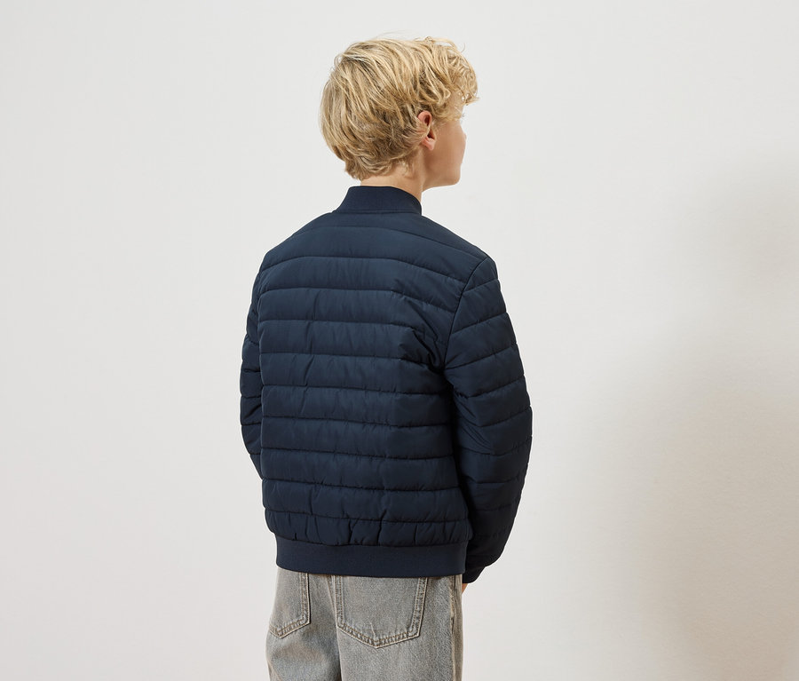 Rückansicht eines blonden Kindes mit marineblauer Steppjacke und grauen Jeans vor einem weißen Hintergrund.