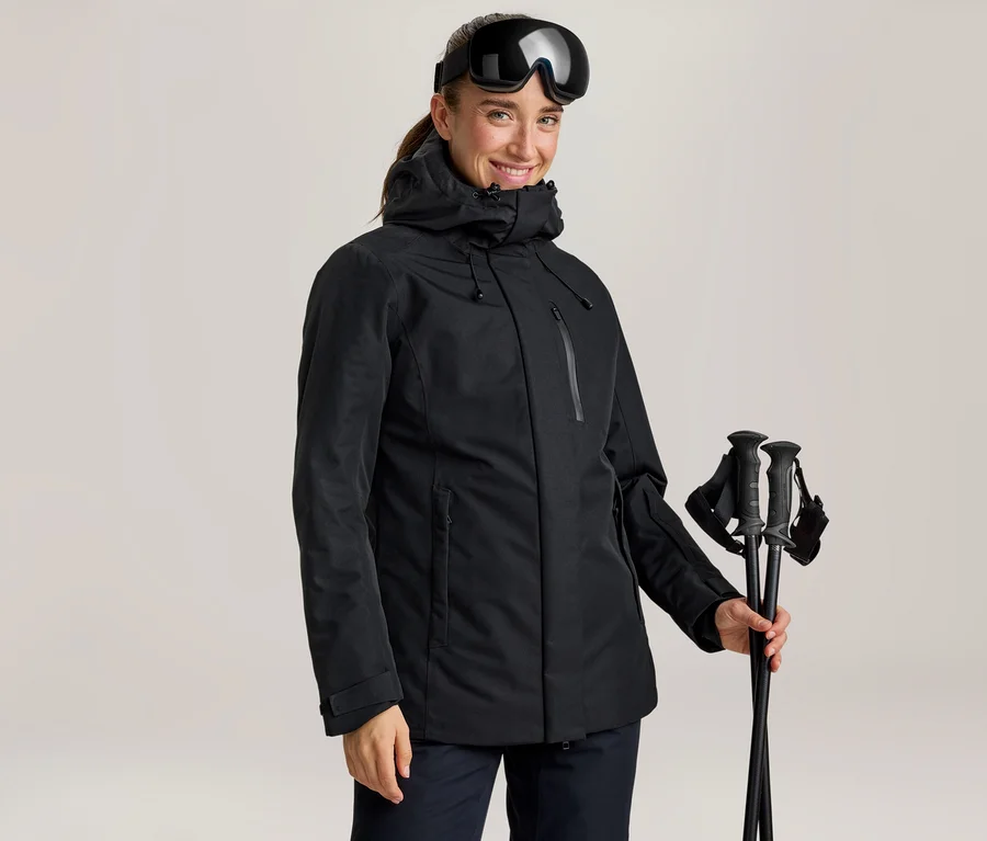 Lächelnde Frau in schwarzer Skijacke mit Skibrille am Kopf, die schwarze Skistöcke hält.