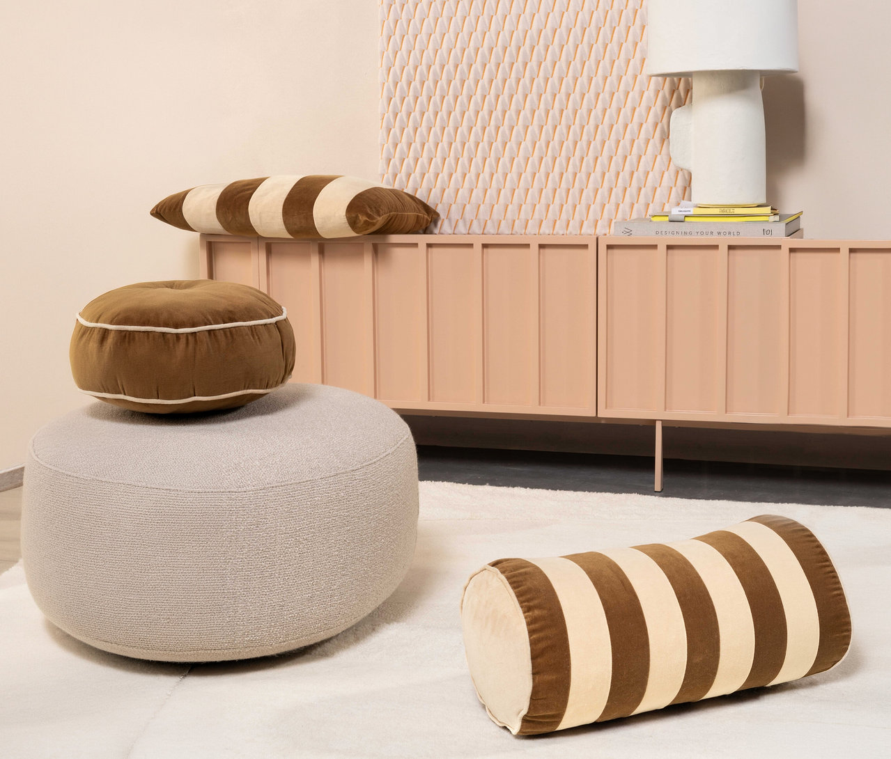 Ein hellbeiger Pouf steht auf einem weißen Teppich, darauf liegt ein rundes, braunes Essenza Samt-Dekokissen »Nino«. Ein zylindrisches, braun-beige gestreiftes Samtkissen liegt auf dem Teppich. Im Hintergrund steht ein pfirsichfarbener Schrank mit einem langen, braun-beige gestreiften Samtkissen, einer weißen Lampe und Büchern.