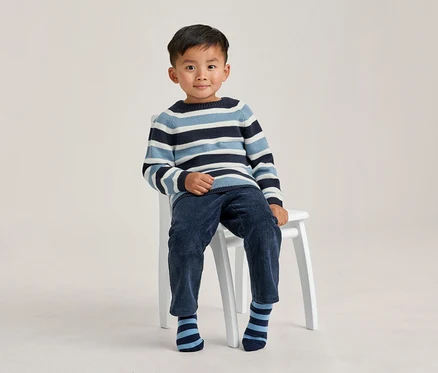 Kleiner Junge sitzt auf einem weißen Hocker, trägt einen blau, hellblau und weiß gestreiften Pullover, dunkelblaue Cordhose und gestreifte Socken, lächelt in die Kamera.