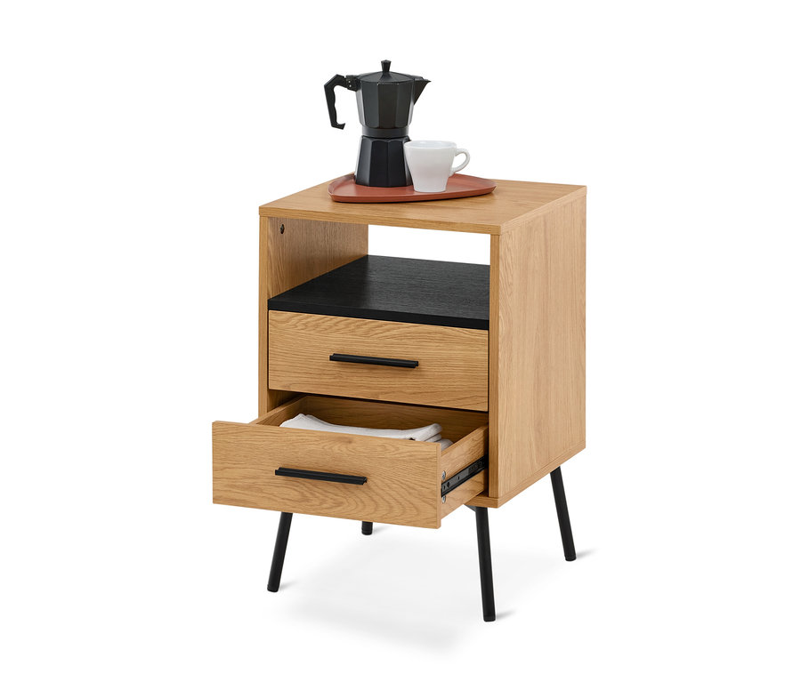 Heller Nachttisch aus Holz mit schwarzen Akzenten und schrägen Beinen. Oben stehen eine schwarze Mokkakanne und eine weiße Tasse auf einem rotbraunen Tablett. Darunter befinden sich ein offenes Fach mit schwarzem Inneren und zwei Schubladen, wobei die untere geöffnet ist und weiße gefaltete Wäsche zeigt.