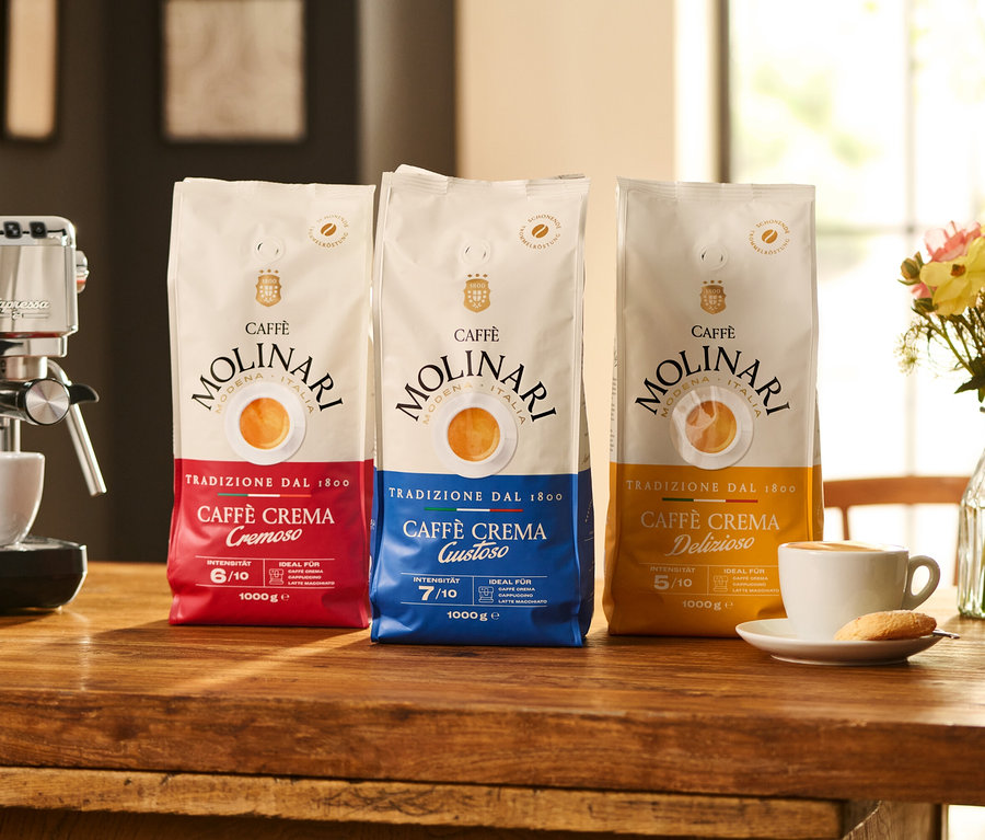 Auf einem Holztisch stehen drei Packungen Caffè Molinari Kaffee neben einer Kaffeemaschine und einer Tasse Kaffee.