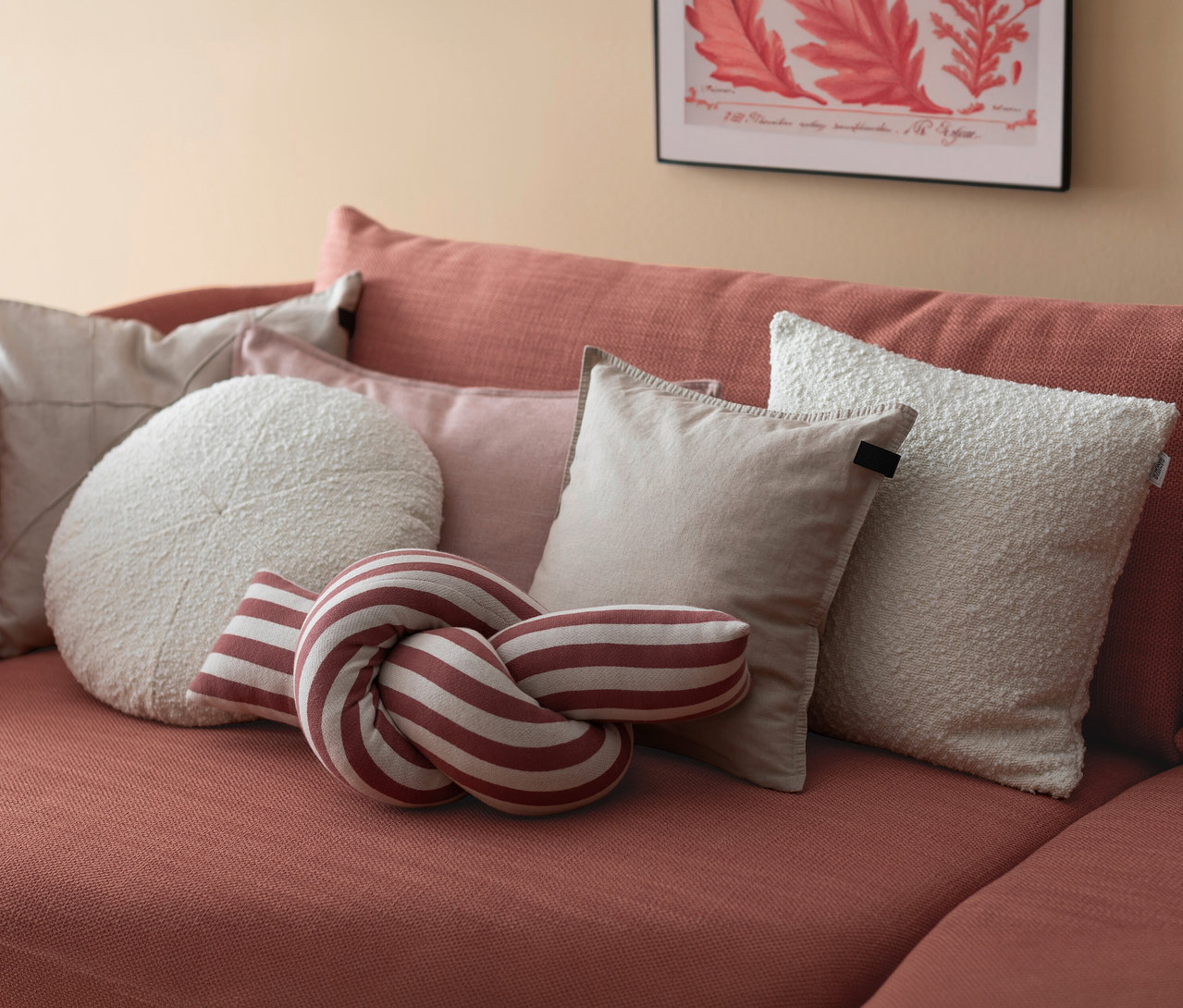 Ein terrakottafarbenes Sofa mit verschiedenen Kissen, darunter ein rot-weiß gestreiftes Knotenkissen »JOLLY«, ein rundes cremefarbenes Kissen und mehrere rechteckige beige und cremefarbene Kissen.