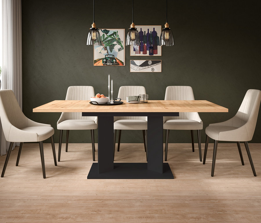 Ein Esszimmer mit einem rechteckigen Esstisch mit heller Holzplatte und schwarzem Fuß, umgeben von sechs beigefarbenen, gepolsterten Stühlen. An der dunkelgrünen Wand hängen Bilder, und über dem Tisch sind drei Pendelleuchten. Auf dem Tisch stehen eine Schale mit Obst und Kerzen. Links ist ein Fenster mit einer Pflanze und einer Gießkanne zu sehen.