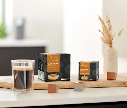 Ein Glas Kaffee, eine 27er- und eine 8er-Packung Qbo Caffè BUNA ENTETA Kapseln sowie zwei einzelne Qbo Kapseln stehen auf einem Holzbrett. Eine Schale mit Keksen und eine Vase mit Trockenblumen sind im Hintergrund.