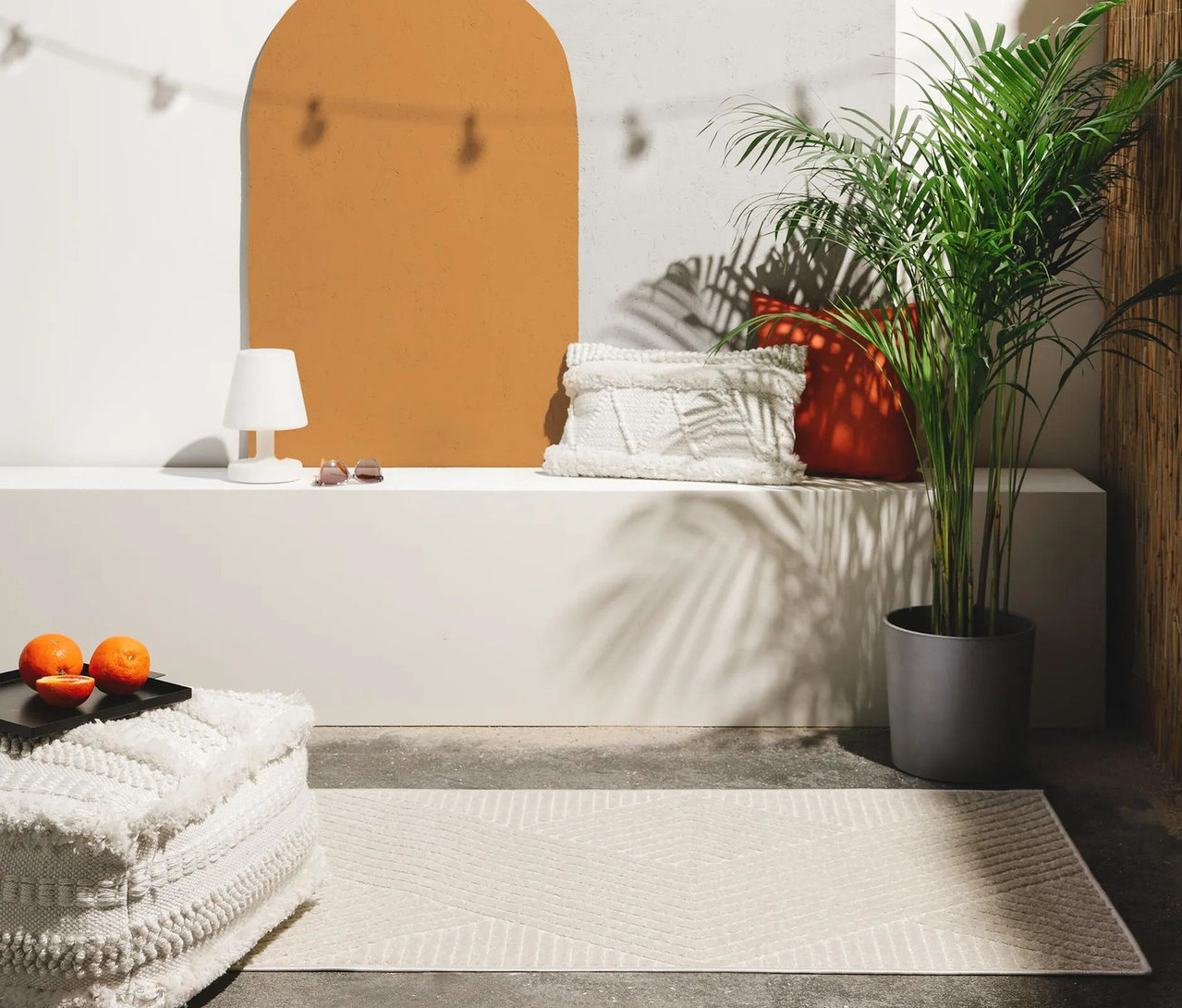 Ein heller benuta Indoor-Outdoor-Läufer »Mars« mit geometrischem Muster liegt vor einer weißen Bank mit Kissen, Lampe und einer Palme.
