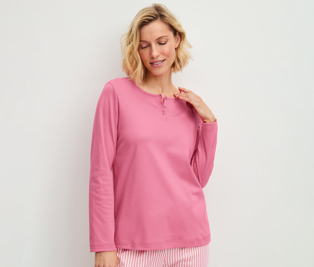 Frau trägt ein rosa Langarmshirt mit Knopfleiste und gestreifte Pyjamahose, blickt lächelnd nach unten.