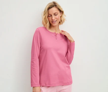 Frau trägt ein rosa Langarmshirt mit Knopfleiste und gestreifte Pyjamahose, blickt lächelnd nach unten.