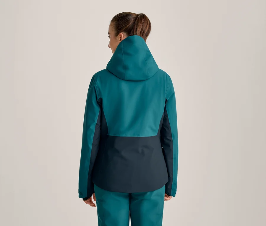 Frau von hinten in einer zweifarbigen Jacke in Petrol und Dunkelblau mit Kapuze und passender Hose, die Haare zum Pferdeschwanz gebunden.