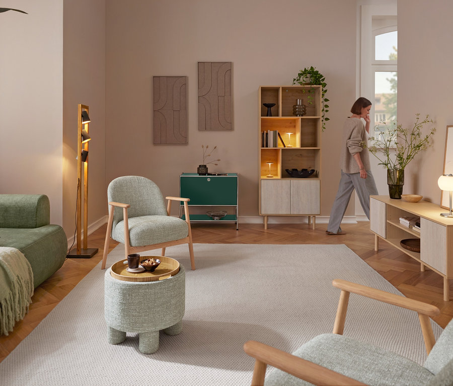 Eine Frau geht durch ein Wohnzimmer mit einem grünen Sofa, zwei Sesseln mit Holzgestell und einem Sitzpouf mit Tablett. Eine Trio Stehleuchte »Bell« steht neben dem Sofa. An der Rückwand stehen ein Sideboard Metall »CN3« und ein Bücherregal in Travertinoptik mit zwei Trio LED-Tischleuchten »Madison«. Rechts steht ein Lowboard in Travertinoptik mit einer Trio LED-Tischleuchte »Madison« und einer Blumenvase.