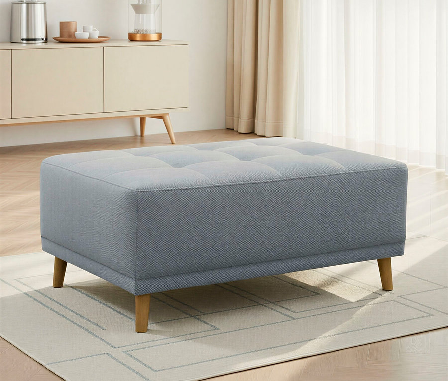 Ein hellblauer, gepolsterter Hocker mit getufteter Oberseite und Holzbeinen steht auf einem hellen Teppich mit geometrischem Muster. Im Hintergrund befindet sich ein Sideboard mit einem Wasserkocher und einem Dekoartikel, neben einem Fenster mit transparenten Vorhängen.