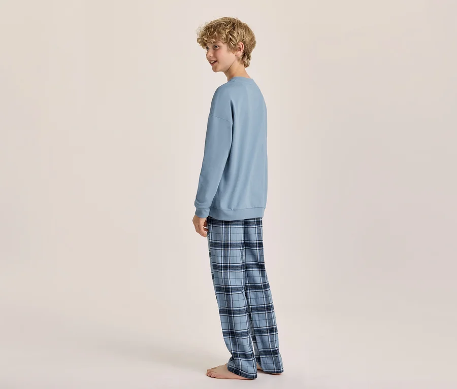 Ein junger Junge mit blonden Locken, der ein hellblaues Langarm-Sweatshirt und eine blau karierte Pyjamahose trägt, steht barfuß und blickt über seine linke Schulter zurück.