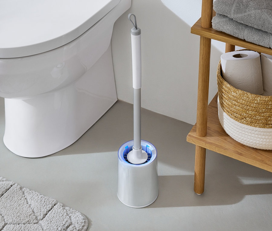 Im Badezimmer steht eine weiße Toilette neben einer WC-Bürste mit blauem Licht und einem Holzregal mit Handtüchern und Toilettenpapier.
