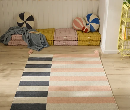 Ein Kinderzimmer mit einem gestreiften Teppich auf Holzboden. Der Teppich hat links vertikale dunkelgraue und cremefarbene Streifen, rechts horizontale hellrosa und cremefarbene Streifen und rosa Quasten. Im Hintergrund eine Sitzecke mit gestapelten rosa und gelben Bodenkissen, einer blauen Decke und runden gestreiften Kissen. Rechts steht ein gelbes Metallregal mit einer Pflanze.