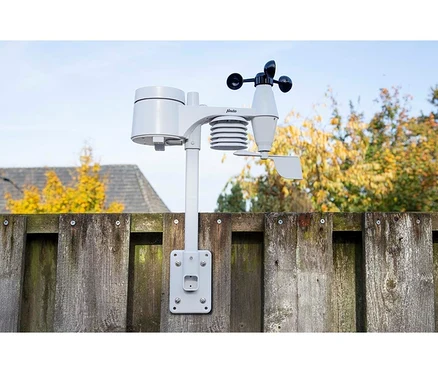 Hellgraue Wetterstation mit Regenmesser, Windfahne und Anemometer an einem verwitterten Holzzaun montiert. Im Hintergrund sind Herbstbäume und ein Hausdach unter klarem Himmel zu sehen.