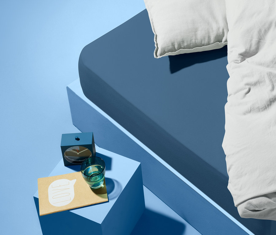 Blaues Bett mit weißem Kissen und Bettdecke. Auf einem blauen Nachttisch neben dem Bett stehen ein Wecker, ein Glas Wasser und ein Notizblock.