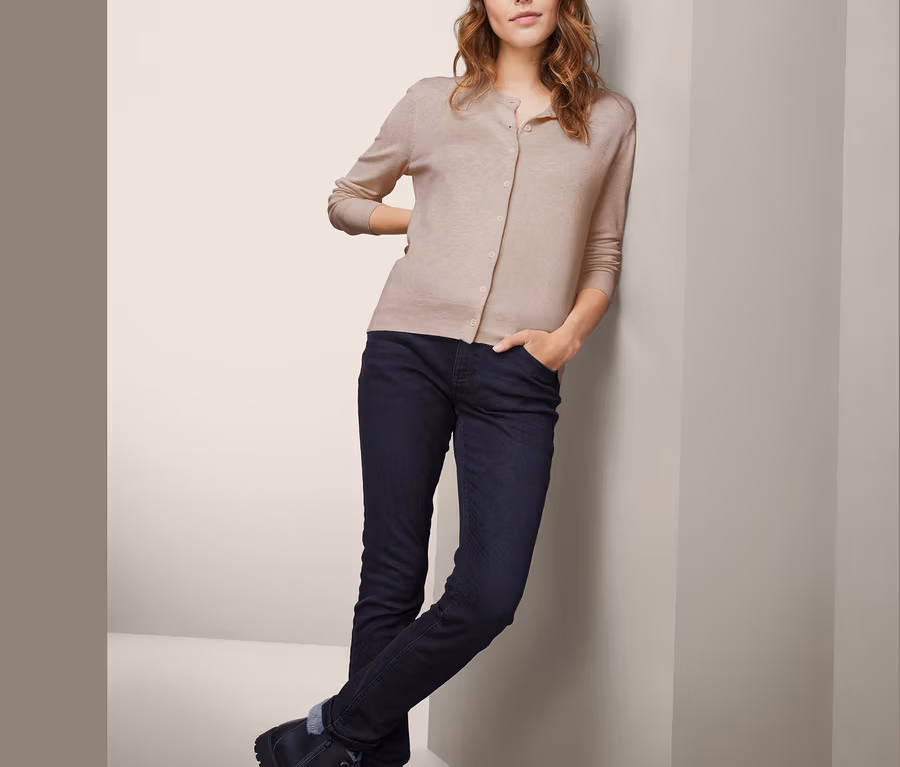 Frau lehnt an Wand, trägt Feinstrickjacke, Jeans und Stiefel.