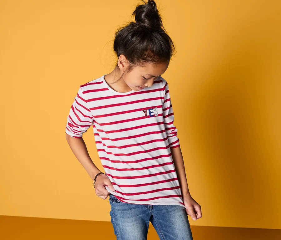 Ein Mädchen trägt ein Langarmshirt mit Wendepailletten-Applikation, blaue Jeans und steht vor einem gelb-orangenen Hintergrund.