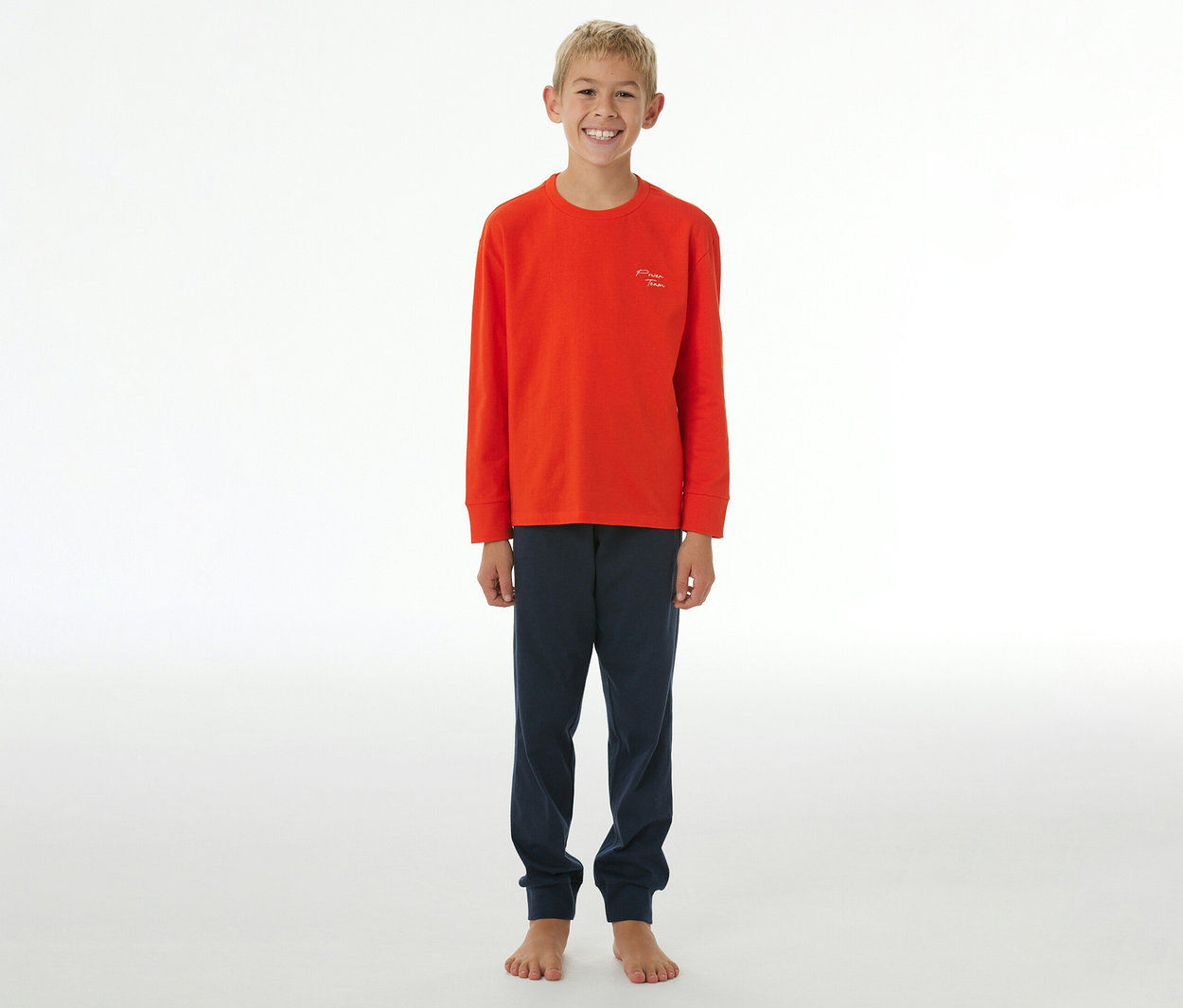 Lächelnder Junge in einem orangefarbenen SCHIESSER Kinder-Pyjama mit langen Ärmeln und dunkelblauer Hose mit Bündchen.