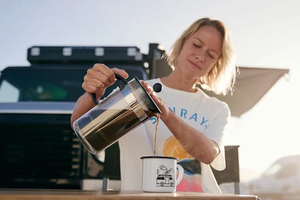 Frau gießt Kaffee aus einer Bodum® Siebstempelkanne in einen Emaille-Kaffeebecher (380 ml). Sie trägt ein T-Shirt mit Print.