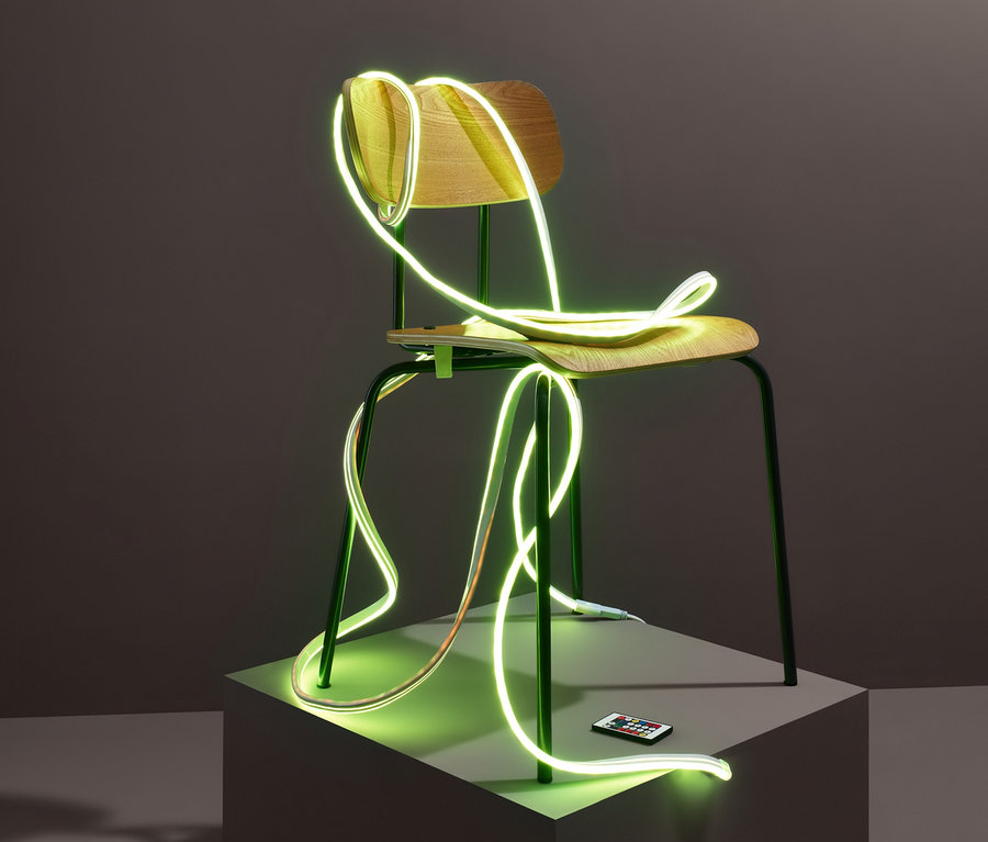 Ein Holzstuhl mit einem LED-Leuchtschlauch mit Lichteffekten, der darum gewickelt ist, steht auf einem Sockel. Auf dem Sockel liegt eine Fernbedienung.