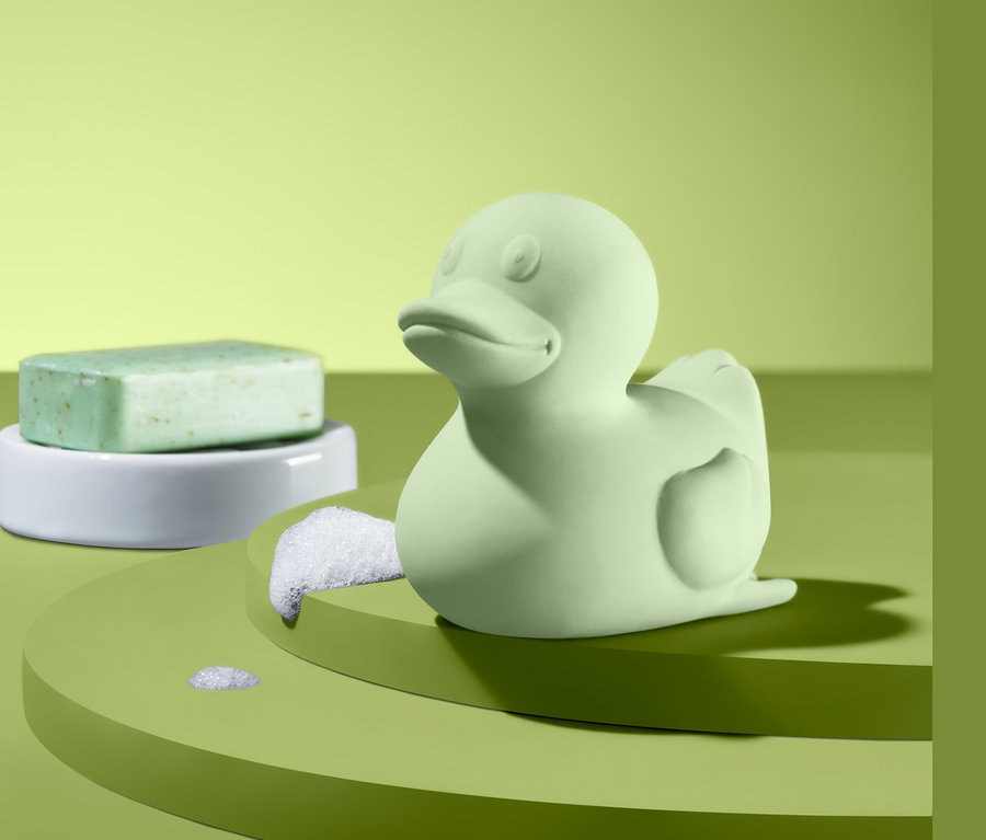Eine kleine, grüne Badeente und ein grünes Seifenstück auf einer weißen Ablage.