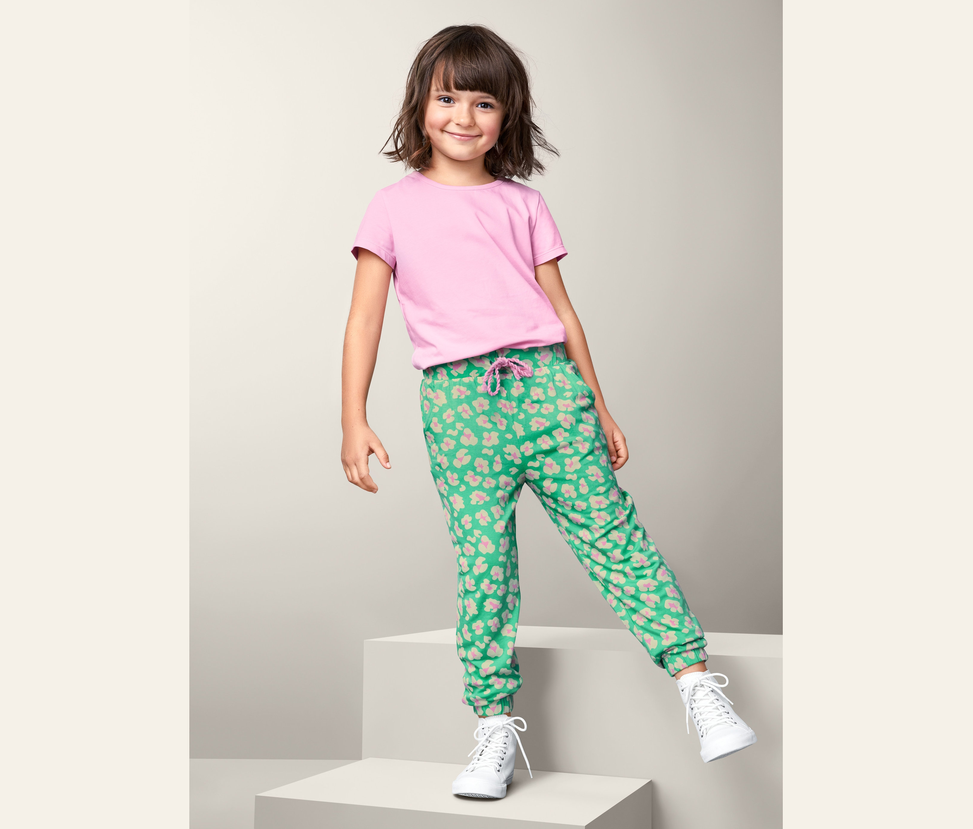 Mädchen in rosa T-Shirt und Jersey-Schlupfhose steht auf einem weißen Würfel.