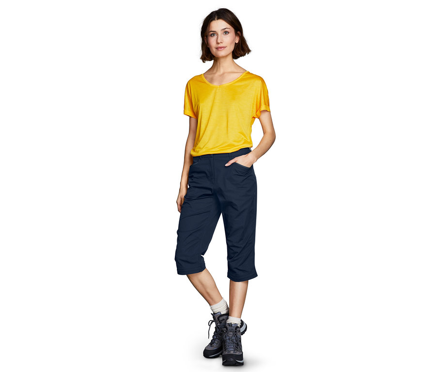 Eine Frau steht in einem gelben T-Shirt und einer marineblauen, abzippbaren Funktionshose.