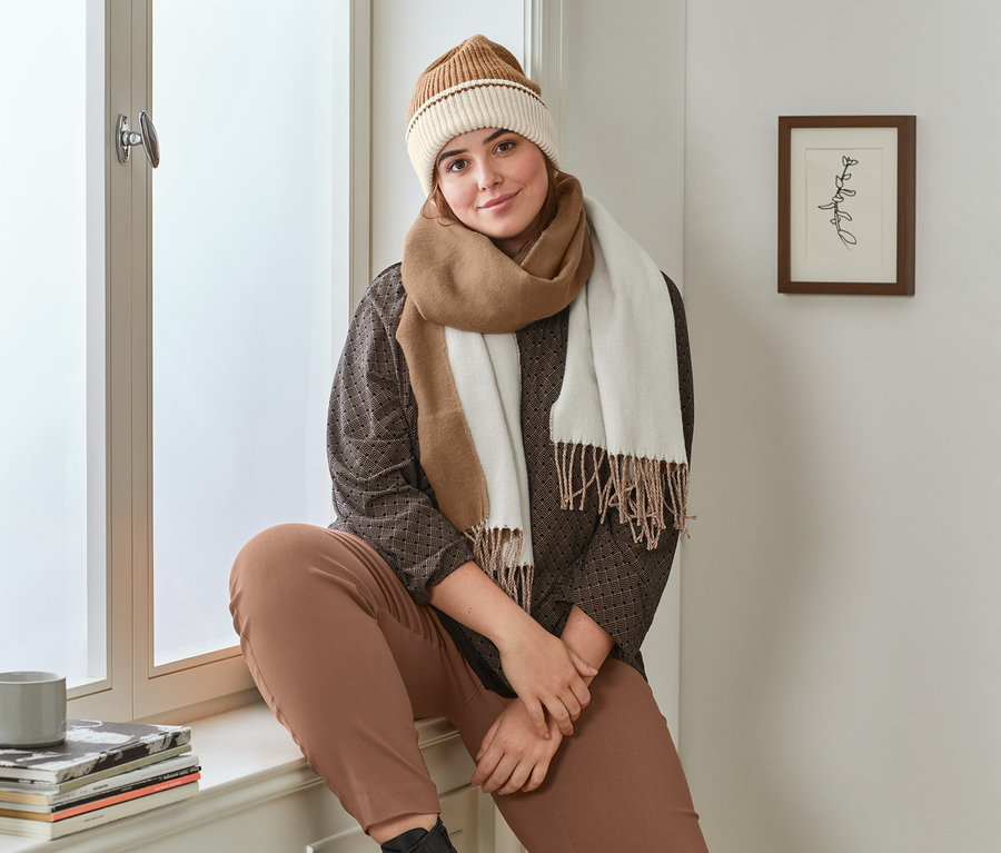 Eine Frau sitzt auf einer Fensterbank und trägt ein bedrucktes schwarzes Blusenshirt, eine cognacfarbene Stretchhose, Leder-Boots, eine creme-cognacfarbene Strickmütze und einen cognac-cremefarbenen Schal mit Fransen.