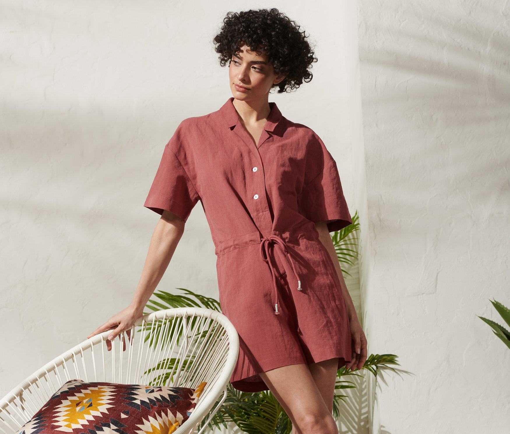 Frau lehnt in kurzem Jumpsuit aus Leinenmix an einem weißen Stuhl.