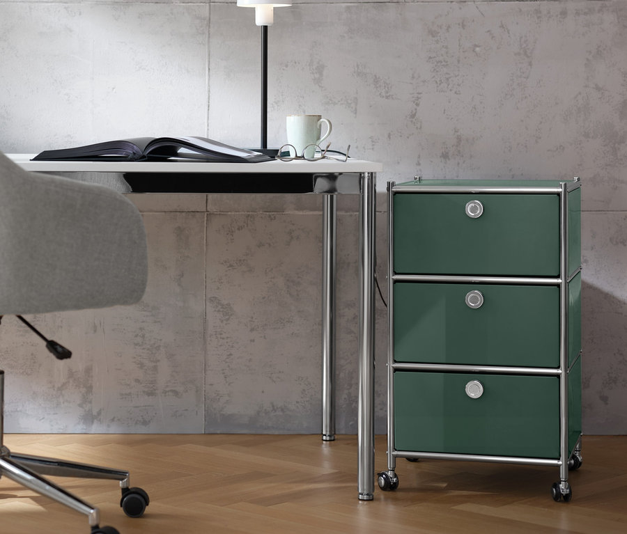 Ein beiger Schreibtischstuhl steht an einem weißen Schreibtisch »CN3«, auf dem ein offenes Buch, eine Tischleuchte aus Metall, eine Brille und eine Tasse stehen. Neben dem Tisch steht ein olivgrüner Rollcontainer »CN3« mit 3 Schubladen.