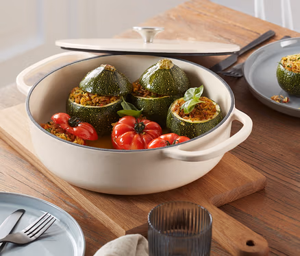 Auf einem Holztisch steht eine cremefarbene Gusseisen-Bräter-Pfanne mit gefüllten Zucchini und Tomaten.