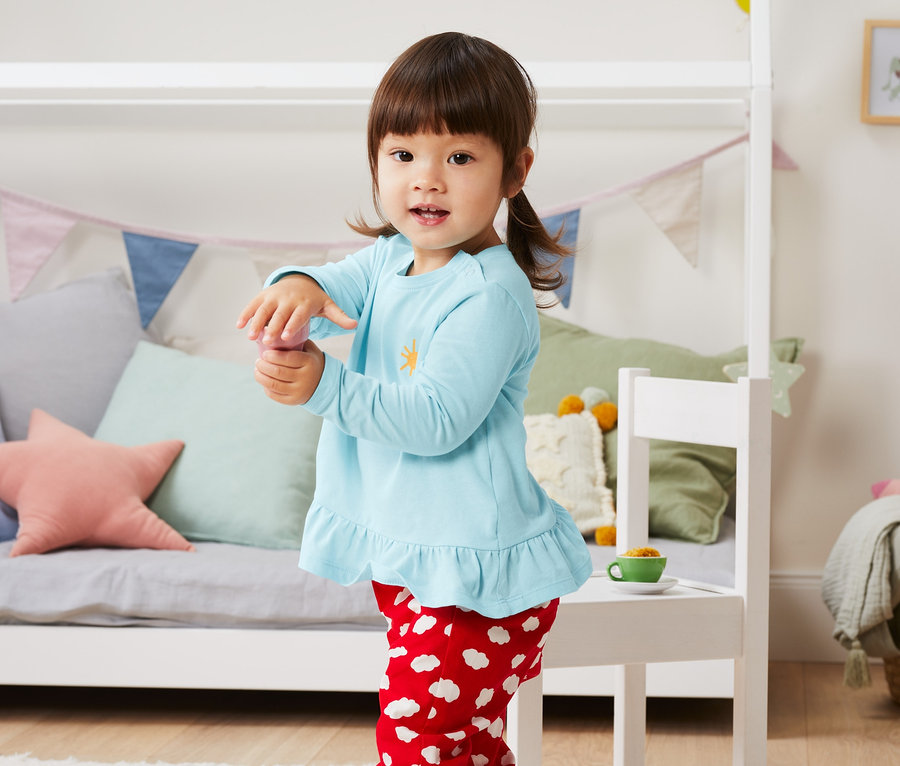 Ein Mädchen steht im Kinderzimmer und trägt ein hellblaues Shirt und rote Hosen mit weißen Wolken. Sie trägt ein 3 Shirts aus Bio-Baumwolle.