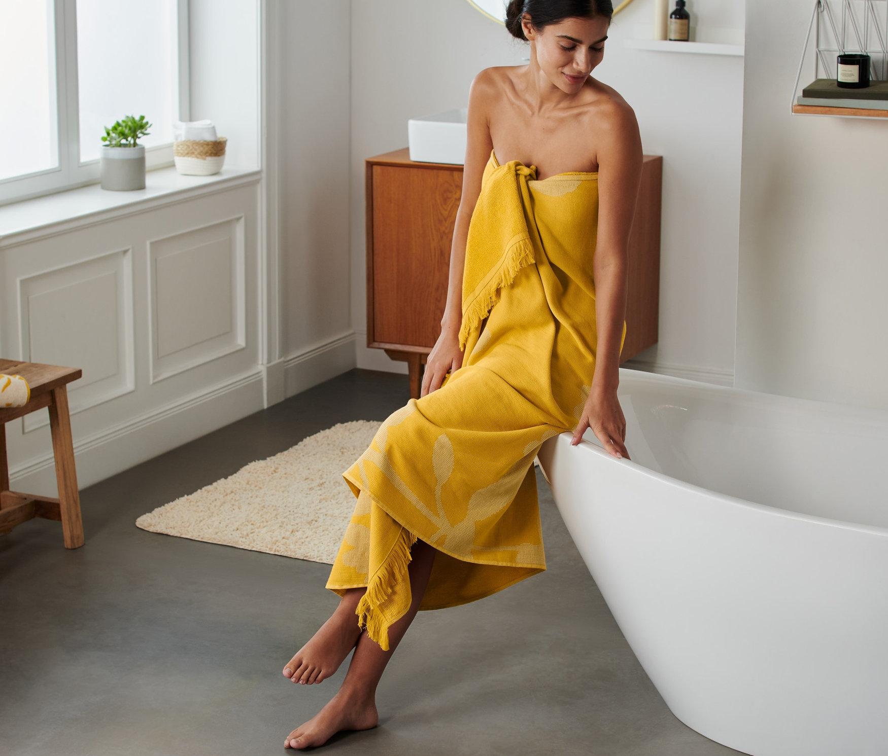 Frau sitzt in gelbes Extragroßes Hamam-Badetuch gehüllt auf dem Badewannenrand.