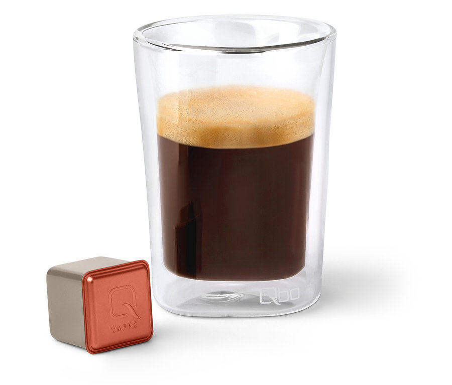 Eine Qbo Caffè TERRENO ALTO Kapsel neben einem Glas Kaffee.