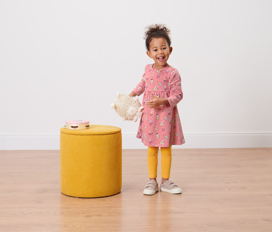 Ein lächelndes kleines Mädchen hält ein Plüschtier und trägt ein Kleinkind-Jerseykleid.