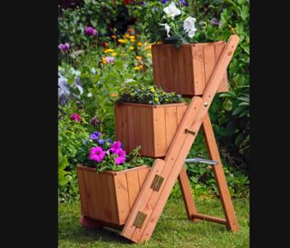 Eine braune Pflanztreppe mit 3 Pflanzkästen steht auf einer Wiese, wobei sich in jedem Kasten Blumen befinden.
