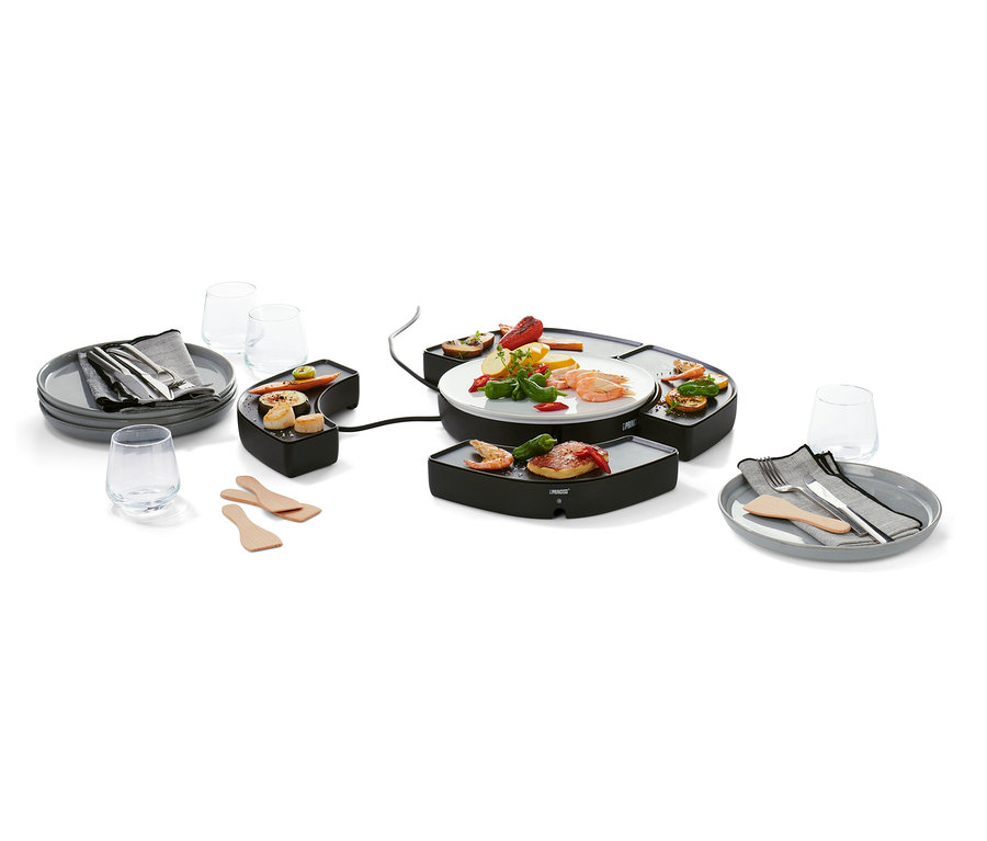 Schwarzer Tischgrill »Dinner4All« mit Speisen auf der Grillfläche und einem Teller. Drumherum Teller mit Besteck, Gläser und Holzspatel.