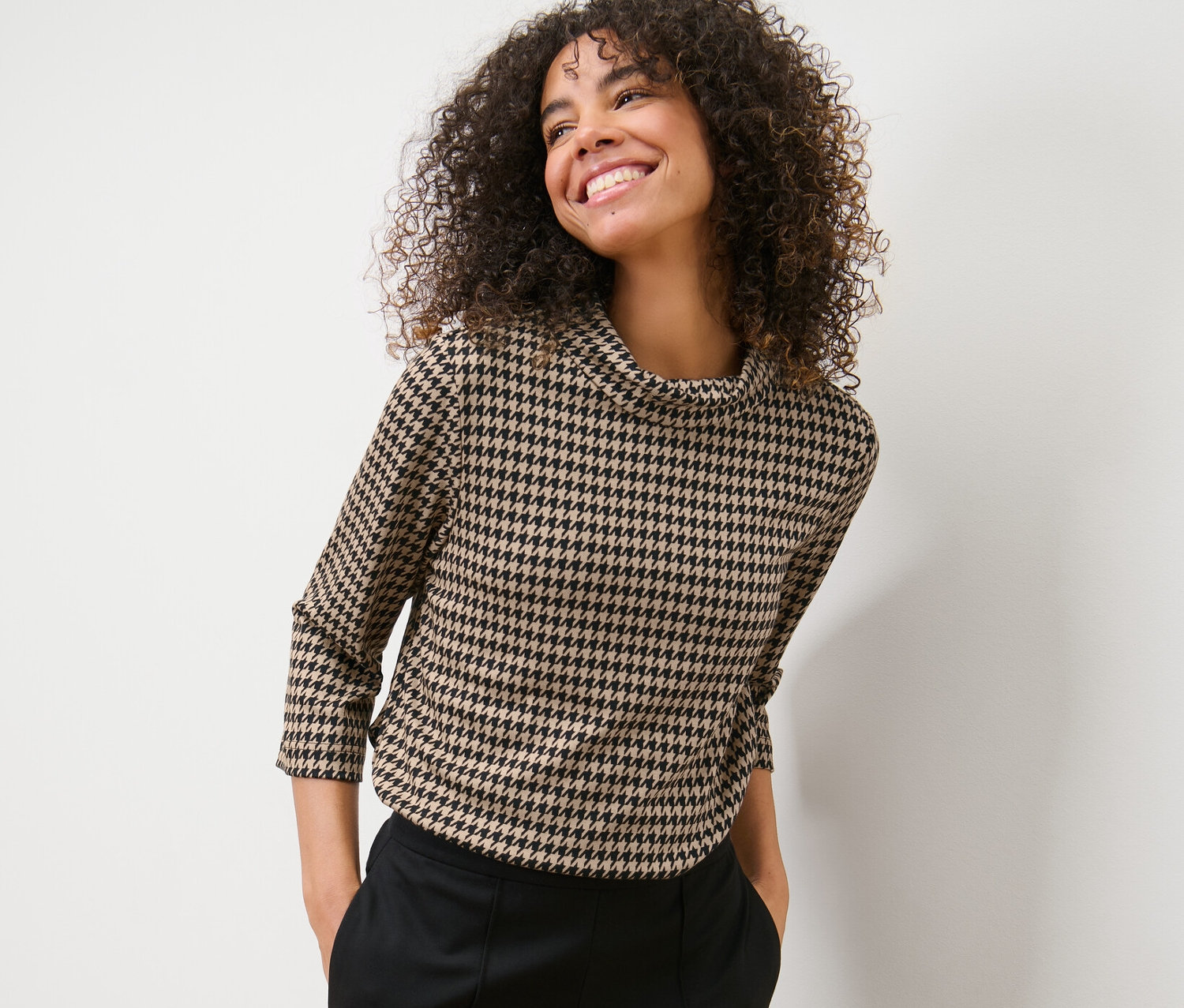 Frau mit lockigem Haar posiert in einem Sweatshirt mit 3/4-Arm, Hahnentrittmuster und einer schwarzen Hose.