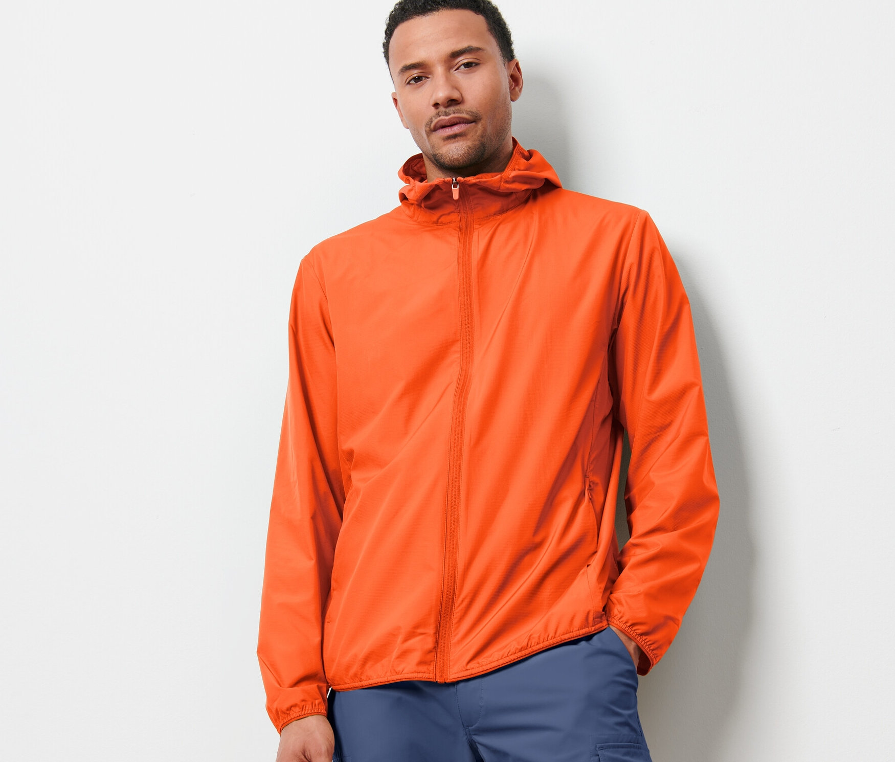 Mann trägt eine orangefarbene, leichte Funktionsjacke »packable« und blaue Hosen und steht vor einer weißen Wand.