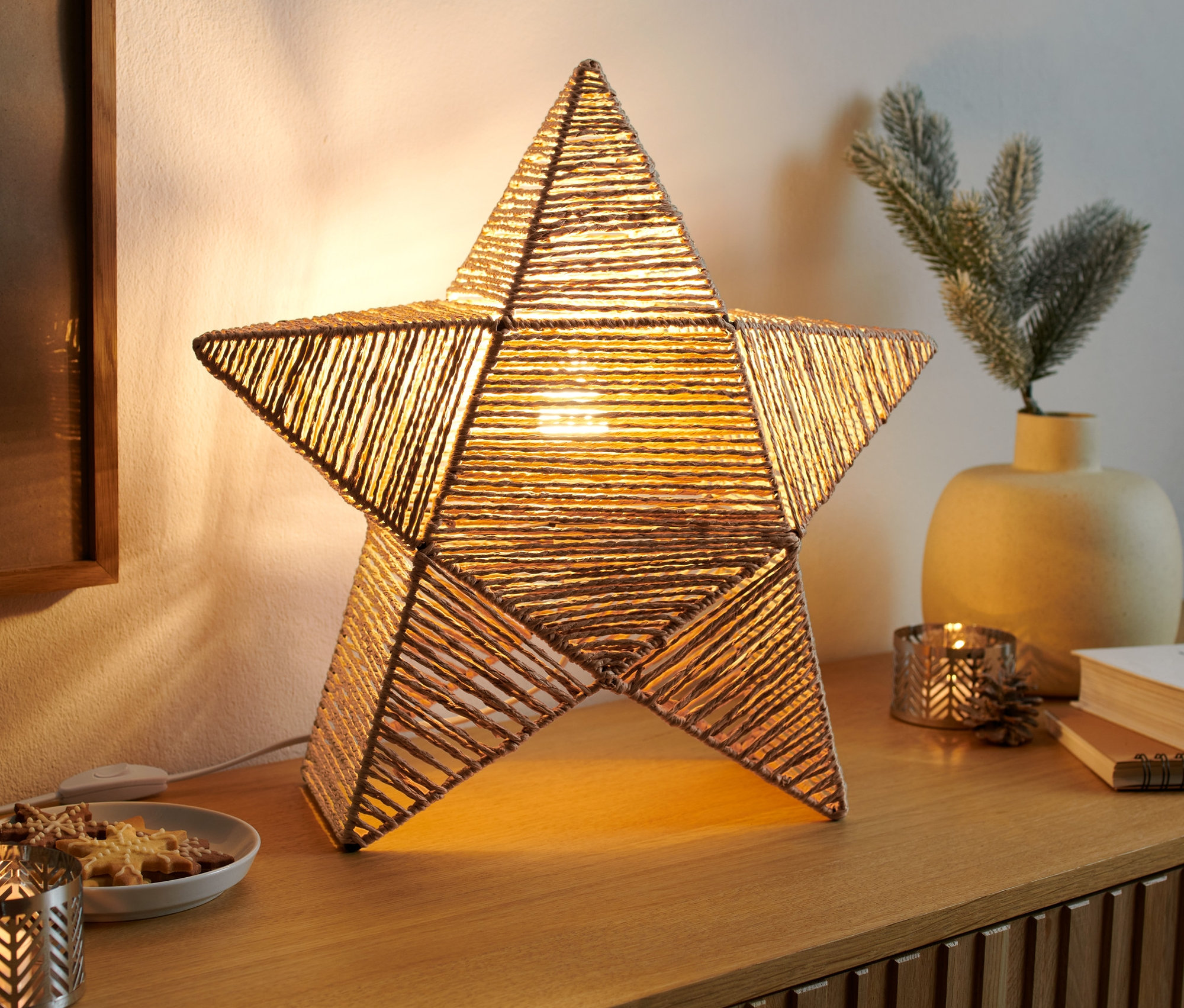 Ein leuchtender LED-Stern aus Naturgeflecht steht auf einer Kommode.