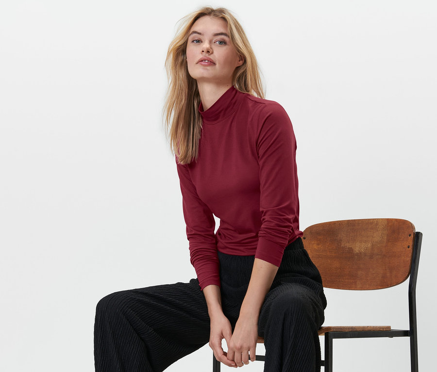 Blonde Frau sitzt auf einem Stuhl und trägt ein rotes Shirt mit Stehkragen und eine schwarze Hose.