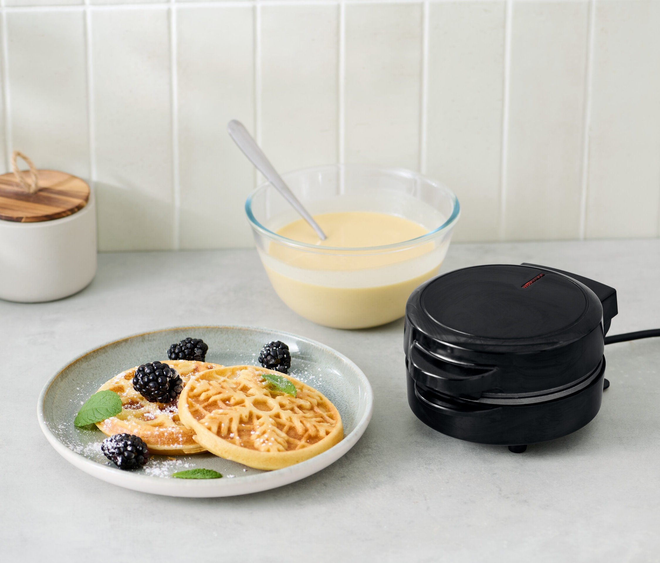 Mini-Waffeleisen neben Teller mit Waffeln, Brombeeren und Minze.