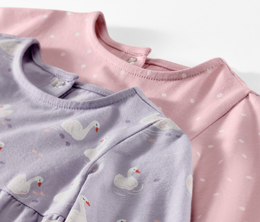 Detailansicht von zwei Baby-Jerseykleidern, eines lila mit Schwanenmuster, das andere rosa mit weißen Punkten.