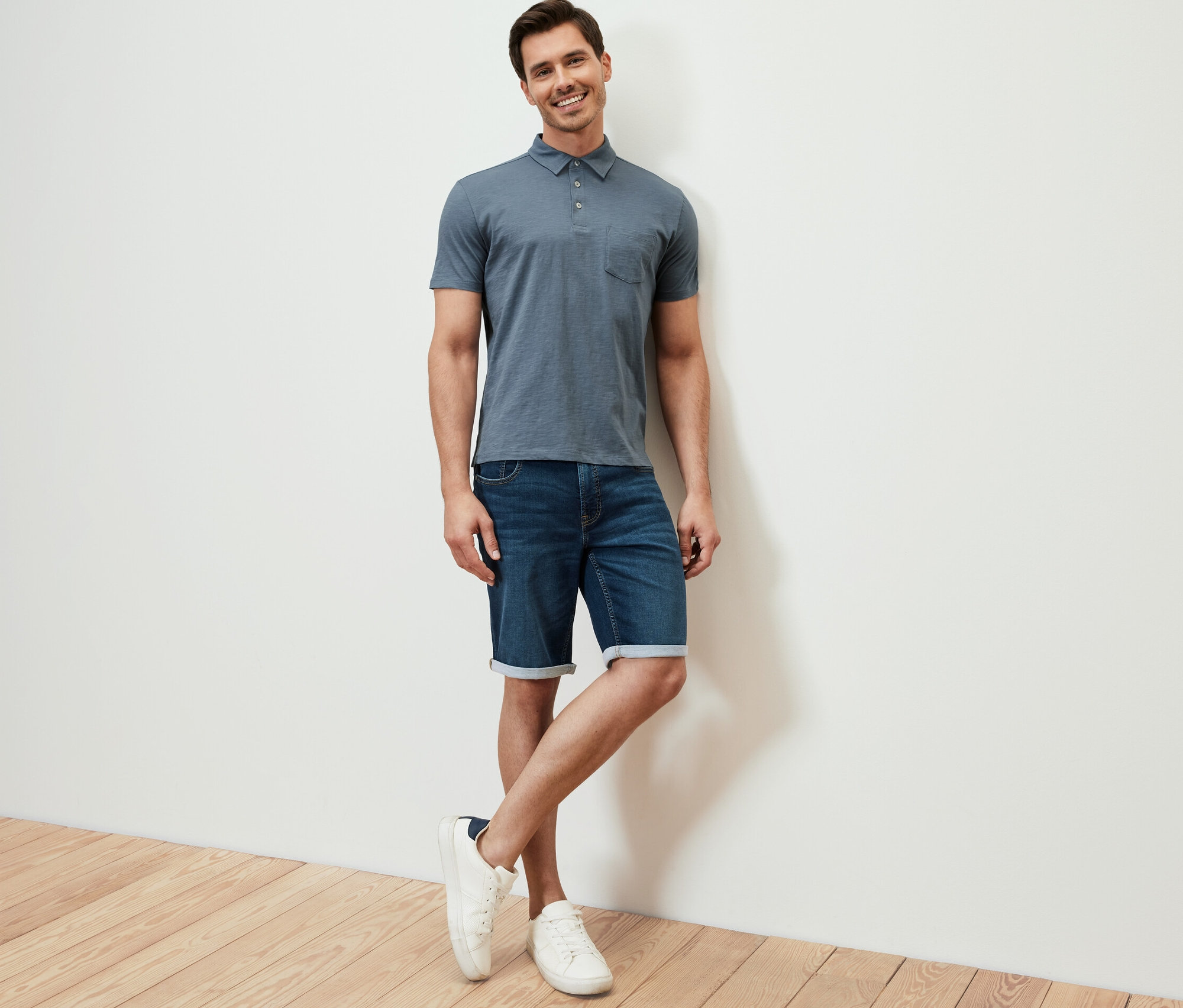 Mann posiert an eine Wand gelehnt und trägt ein hellblaues Jersey-Poloshirt, Jeansshorts und weiße Turnschuhe.
